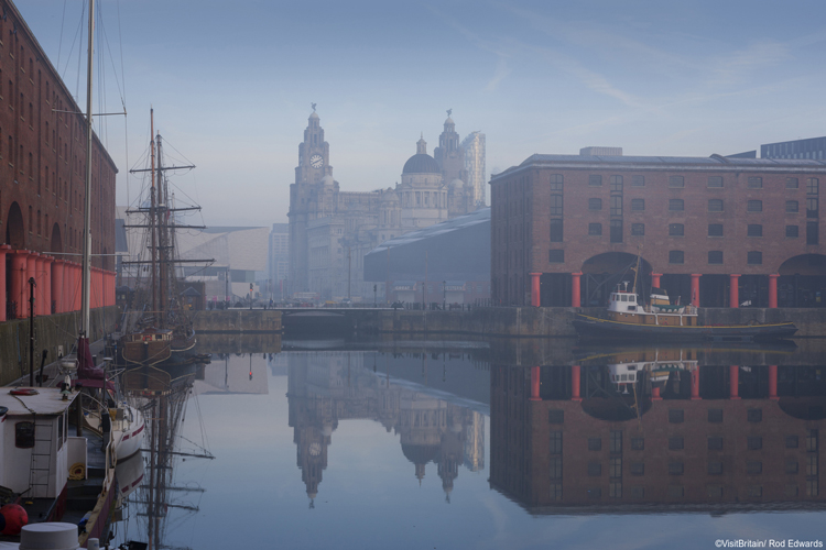 Maritime Albert Dock on Liverpool waterfront with the Three Graces historic buildings. The Liverpool world heritage site. Port of Liverpool building, the Cunard Building and the Liver building. A historic ship moored. Mist rising from the water. Credit Visit Britain/Rod Edwards