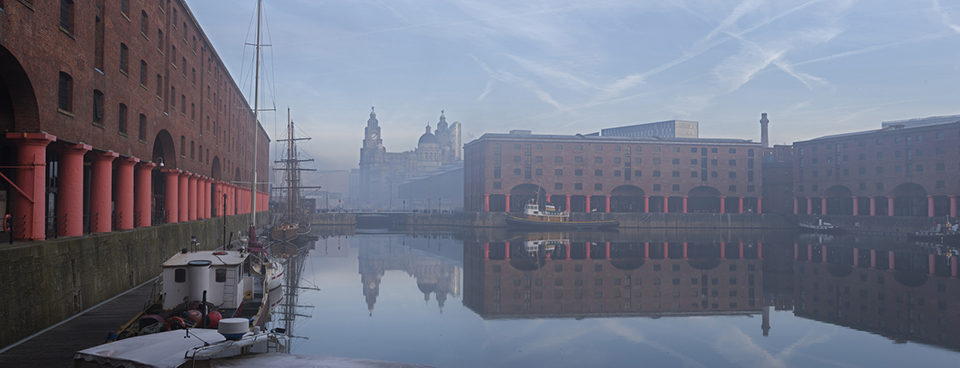 Maritime Albert Dock on Liverpool waterfront with the Three Graces historic buildings. The Liverpool world heritage site. Port of Liverpool building, the Cunard Building and the Liver building. A historic ship moored. Mist rising from the water.